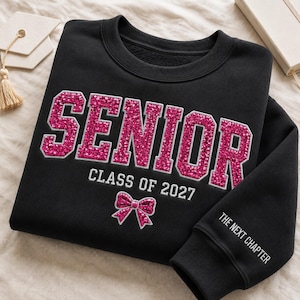 May include: A black crewneck sweatshirt featuring the word SENIOR in large, pink glittery varsity-style letters. Below, the text reads CLASS OF 2027 with a small pink glittery bow icon. The right sleeve cuff displays the text THE NEXT CHAPTER.