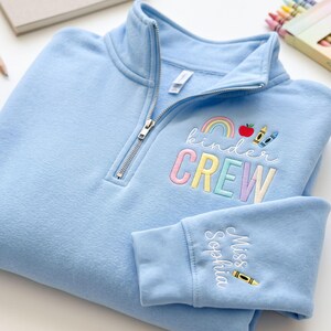 May include: A light blue quarter-zip sweatshirt featuring embroidered text that reads "kinder CREW" in pastel colors, accompanied by a rainbow, apple, and crayon icon. The sleeve is embroidered with the name "Miss Sophia" and a yellow crayon.