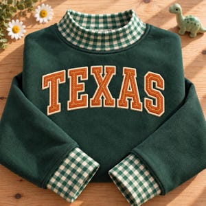 Op de afbeelding: Een donkergroen sweatshirt met lange mouwen en het woord TEXAS in oranje geborduurde blokletters op de borst. Het kledingstuk heeft een opstaande kraag en mouwboorden met een groen-wit vichy-ruitpatroon.