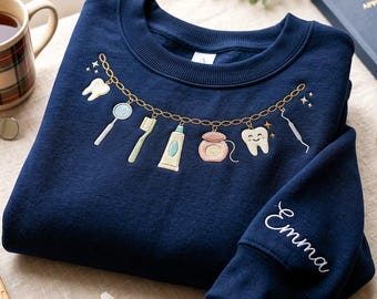 Puede incluir: Una sudadera azul marino de cuello redondo con un diseño bordado de cadena dorada en el pecho con dijes de temática dental que incluyen un diente, espejo, cepillo, pasta, hilo dental y un diente sonriente. El nombre Emma está bordado en la manga.