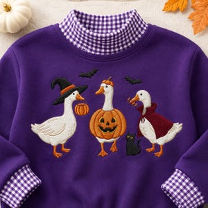 May include: A purple sweatshirt featuring a mock neck with a purple and white gingham pattern. The front displays an embroidered design of three white geese in Halloween costumes, including a witch, a pumpkin, and a vampire, with a black cat.