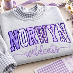 May include: A light grey sweatshirt featuring a mock neck collar with a grey and white gingham pattern. The front displays the embroidered text NORWYN in purple block letters with floral and gingham patterns, and wildcats in purple script.