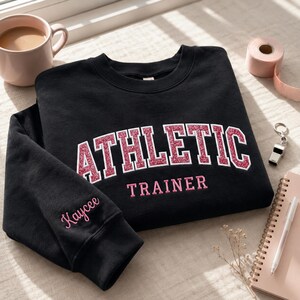 May include: A black crewneck sweatshirt featuring the word ATHLETIC in pink glittery block letters with a white outline, and the word TRAINER in solid pink letters below. The name Kaycee is embroidered in pink script on the left sleeve cuff.