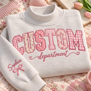 Op de afbeelding: Een lichtgrijs sweatshirt met een opstaande kraag en het woord CUSTOM in grote roze letters met patronen. Daaronder is het woord department in roze cursief geborduurd met kleine hartjes. De mouw toont de naam Sophia met een roze strik.