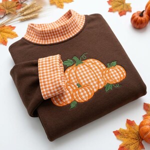 Op de afbeelding: Een donkerbruin sweatshirt met lange mouwen, een opstaande kraag en mouwboorden in een oranje en wit vichy-ruitpatroon. De voorzijde van het kledingstuk toont een geborduurd ontwerp van drie pompoenen in hetzelfde oranje en witte vichy-patroon.