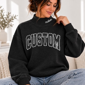 May include: A black long-sleeve sweatshirt with a mock neck collar. The word "custom" is embroidered in white script on the neck. The chest features the word "CUSTOM" in a large, arched, white-dotted collegiate font with a gray shadow effect.