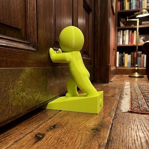 Op de afbeelding: Een fel neongele deurstopper in de vorm van een gestileerd menselijk figuur dat tegen een houten deur duwt. Het figuur staat in een voorovergebogen houding, met de handen plat tegen de deur en de voeten op de wigvormige basis.