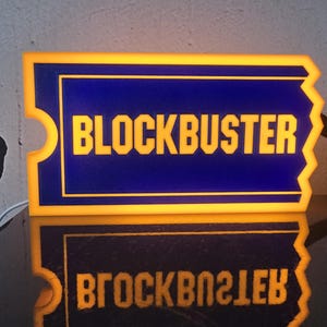 May include: A rectangular, ticket-shaped illuminated sign featuring a deep blue background with a bright yellow border and the word BLOCKBUSTER in bold, yellow, capitalized letters. The sign emits a warm glow from its edges and center.