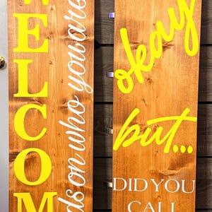 May include: Two tall wooden porch signs with natural wood grain. The left sign features yellow text reading WELCOME ish and white text reading depends on who you are. The right sign features yellow text reading okay but and white text reading DID YOU CALL FIRST. Both signs are topped with yellow flowers and black and white plaid bows.