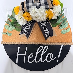 May include: A round wooden door sign featuring a natural wood top half and a black bottom half with the white text Hello!. It is decorated with a black and white buffalo check bow, yellow and white flowers, and green eucalyptus leaves.