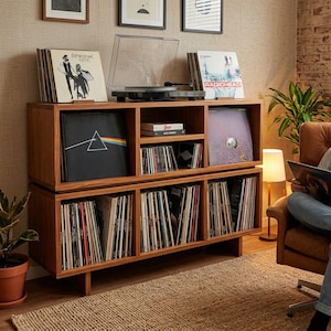 May include: A two-tier wooden media console featuring open shelving filled with vinyl record albums and compact discs. A turntable sits on the top surface, flanked by record sleeves including Fleetwood Mac and Radiohead. The unit has a natural wood finish.