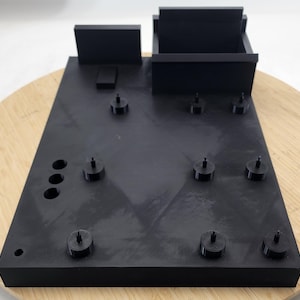 May include: A matte black rectangular organizer tray featuring a raised back wall, a square storage compartment, and multiple small cylindrical pegs arranged across the flat surface. The tray also includes three circular holes on the left side.