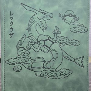 May include: A sage green zippered binder featuring a black line art illustration of the serpentine creature Rayquaza surrounded by stylised clouds and a Poké Ball. The Japanese text "レックウザ" is printed vertically on the left side of the cover.
