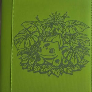May include: A lime green binder featuring a dark green line art illustration of a Bulbasaur character surrounded by tropical monstera leaves and foliage. The cover has a textured, fabric-like finish with a vertical stitched seam on the left side.