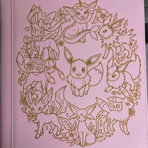 May include: A light pink binder featuring a gold-colored line art illustration of Eevee and its various evolutions arranged in a circular pattern. The design includes floral accents, leaves, and small hearts surrounding the characters on the cover.