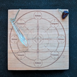Könnte beinhalten: Ein quadratisches Holzbrett mit einem lasergravierten kreisförmigen Diagramm mit den Wörtern Initiate, Integrate und Release. Ein klares, facettiertes Kristallpendel hängt an einer silbernen Kette links, und eine dunkelbraune Steinperle ist rechts befestigt.