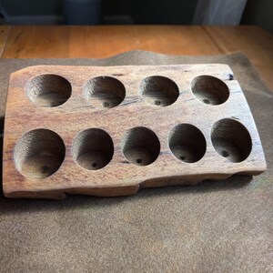 Può includere: Un vassoio rettangolare in legno con bordo naturale e nove scomparti circolari incassati disposti su due file. Il legno presenta una venatura visibile con toni marroni e una consistenza superficiale liscia e rifinita su tutto l'oggetto.