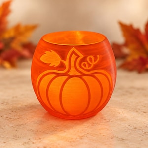 Può includere: Un portacandele rotondo in vetro arancione con un disegno di zucca intagliato sul davanti. Il vetro ha una finitura satinata e testurizzata che brilla di una luce calda, mettendo in risalto le linee incise della zucca, del picciolo e di una foglia.