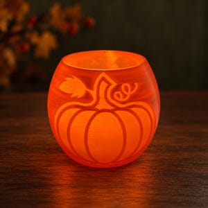 Può includere: Un portacandele sferico in vetro arancione con un design a zucca intagliato sulla parte anteriore. Il materiale traslucido brilla di una luce calda, mettendo in risalto i dettagli incisi del gambo, della foglia e del viticcio.