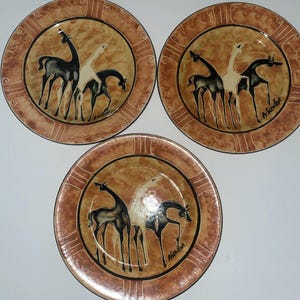 May include: Three round ceramic plates featuring a brown and tan mottled background with a central illustration of three giraffes in black and white. Each plate displays the signature A. Servillon and has a wide, patterned border in shades of brown.