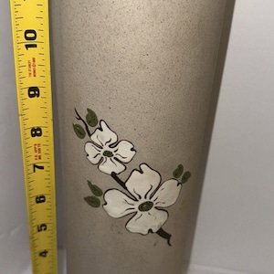 May include: A tall, cylindrical, speckled beige ceramic vase featuring a painted branch with two white dogwood flowers and green leaves. A yellow measuring tape is positioned vertically next to the vase, indicating a height of 30.5 centimetres.