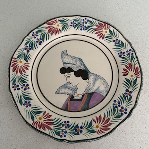 May include: A round ceramic plate with a cream-colored center featuring a profile illustration of a person wearing a decorative headpiece and purple garment. The rim is decorated with a repeating pattern of red flowers and green leaves.