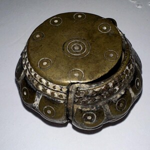 May include: A round, dark bronze-toned metal container with a removable lid. The surface features embossed circular patterns with central dots, and a textured, light-colored band encircles the middle section of the object's exterior body.