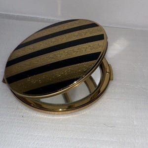 May include: A round compact mirror with a gold-tone metal frame. The lid features alternating horizontal stripes of black and textured gold with an intricate floral pattern. The mirror is partially open, revealing a reflective glass surface inside.