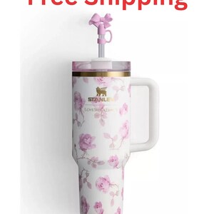 May include: A white tumbler with a pink floral pattern, a gold-tone Stanley logo, and the text LOVESHACKFANCY. It features a side handle, a clear lid, and a metal straw topped with a small pink bow accessory. The design includes pink roses.