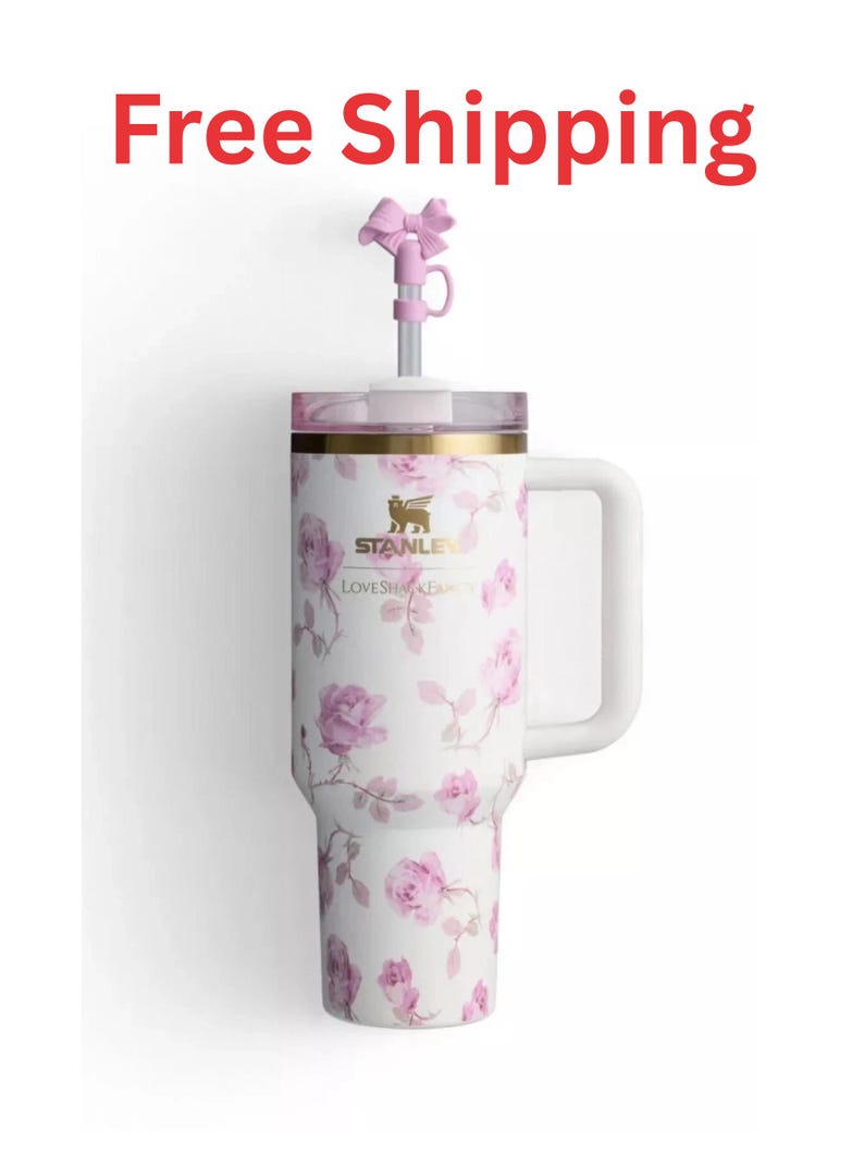 May include: A white tumbler with a pink floral pattern, a gold-tone Stanley logo, and the text LOVESHACKFANCY. It features a side handle, a clear lid, and a metal straw topped with a small pink bow accessory. The design includes pink roses.