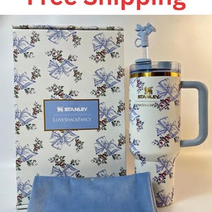 May include: A white Stanley tumbler with a blue bow and floral pattern, featuring a matching straw topper, a rectangular box with the same print and Stanley LoveShackFancy branding, and a folded light blue velvet fabric accessory in front.