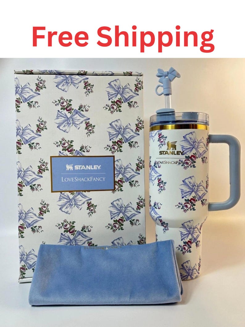 May include: A white Stanley tumbler with a blue bow and floral pattern, featuring a matching straw topper, a rectangular box with the same print and Stanley LoveShackFancy branding, and a folded light blue velvet fabric accessory in front.