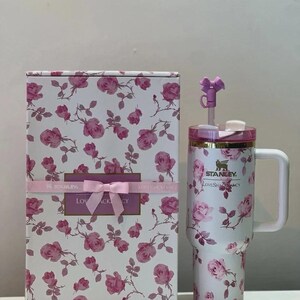 May include: A white Stanley tumbler with a pink rose pattern, a handle, a clear lid, and a purple bow straw topper, displayed next to a matching rectangular box featuring the same floral print, a pink ribbon, and the text LoveShackFancy.
