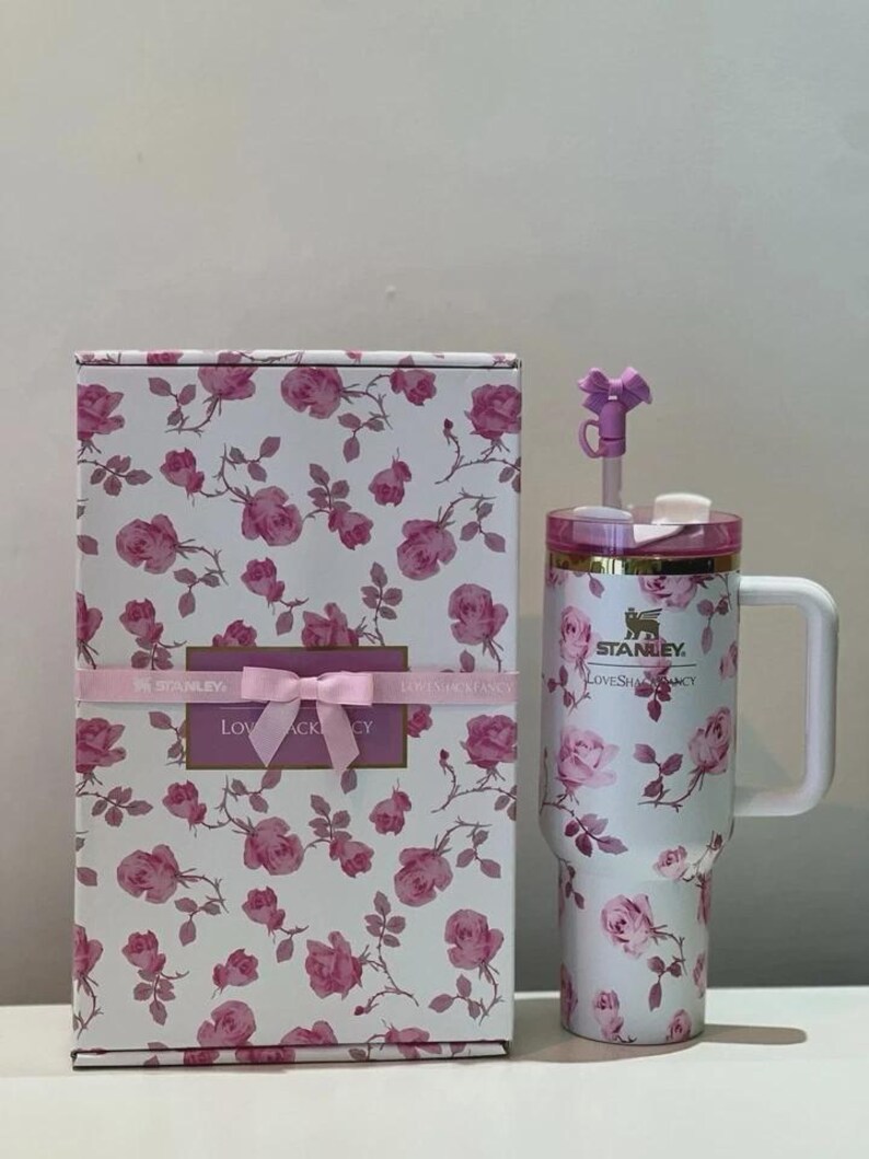May include: A white Stanley tumbler with a pink rose pattern, a handle, a clear lid, and a purple bow straw topper, displayed next to a matching rectangular box featuring the same floral print, a pink ribbon, and the text LoveShackFancy.