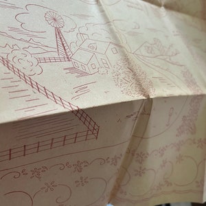 May include: A vintage embroidery pattern featuring a farmhouse with a windmill, a fence, and a flower border. The pattern is printed on a beige paper.