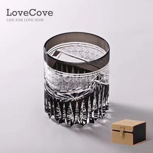 May include: A smoke-coloured crystal whisky glass featuring intricate diamond-cut patterns and vertical fluting at the base. The glass is accompanied by a small, square, tan and black storage box with a central snap closure and the text LoveCove.