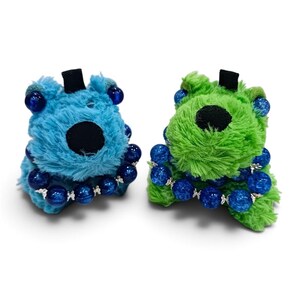 Può includere: Due figure in peluche, una blu e una verde, ciascuna dotata di un naso nero, una piccola linguetta in tessuto nero sulla parte superiore e un braccialetto composto da perline sferiche testurizzate blu scuro con distanziatori argentati.
