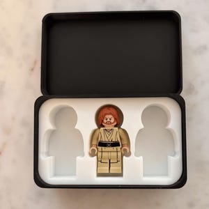 May include: A plastic minifigure with reddish-brown hair and a beard, wearing a tan tunic with a black belt, positioned in the centre slot of a black rectangular storage case with a white foam insert featuring three human-shaped cutouts.