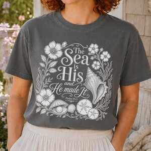 May include: A charcoal gray short-sleeve t-shirt featuring a white line-art graphic of flowers, seashells, and a pearl. The text reads: The Sea is His and He made It Psalm 95:5. The design is centered on the chest of the crew neck garment.