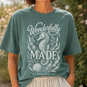 Könnte beinhalten: Ein salbeigrünes T-Shirt mit einem weißen Grafikdruck eines Seepferdchens, Seetang und einer Muschel. Der Text lautet Wonderfully MADE und Psalm 139:14. Das T-Shirt hat einen Rundhalsausschnitt und kurze Ärmel mit einer detaillierten Illustration.