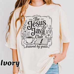 May include: An ivory t-shirt featuring a black graphic print with the text The Jesus & Java Club brewed by grace. The design includes a coffee cup on a saucer, an open book, and floral accents enclosed within a decorative rectangular border.
