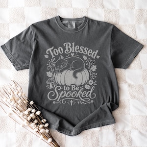 May include: A charcoal gray short-sleeve t-shirt featuring a light gray graphic of a cat sleeping on a pumpkin. The text reads Too Blessed to Be Spooked, surrounded by illustrations of leaves, flowers, a small mushroom, and a tiny cross symbol.