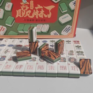 May include: A Mahjong travel set featuring white tiles with green and brown tiger-stripe patterns on the backs. The tiles display various colorful symbols, Chinese characters, and the word JOKER. The set includes a box with red and green graphics.