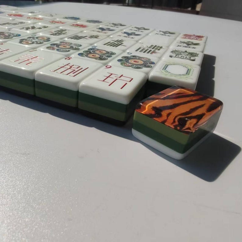 May include: A set of rectangular Mahjong tiles with white faces featuring various red, black, and green symbols, numbers, and text including Joker and RAM. Each tile has a layered green and white base, with one tile displaying a tiger stripe pattern.