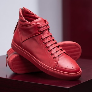 May include: A pair of vibrant red high-top sneakers featuring a smooth leather upper, matching red laces, and a perforated toe box. The design includes a padded collar, a rear zipper closure, and a solid red rubber sole with a textured tread pattern.