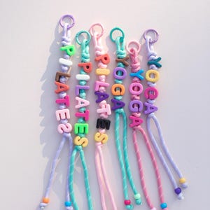 May include: Colorful beaded keychains spelling out "PILATES" and "YOGA". Each keychain features a different color scheme for the beads and cord, with a metal ring at the top for attaching keys. The keychains are a fun accessory.