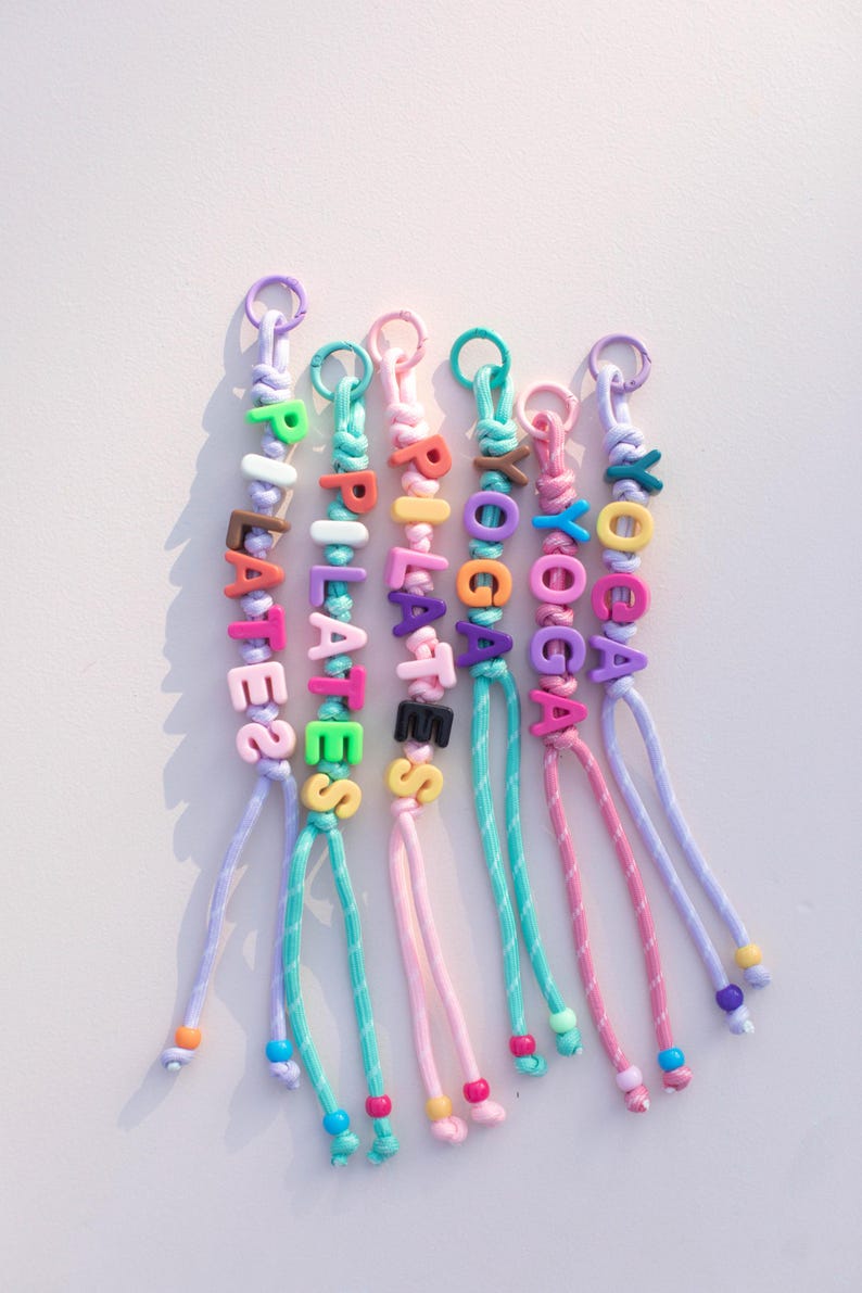 May include: Colorful beaded keychains spelling out "PILATES" and "YOGA". Each keychain features a different color scheme for the beads and cord, with a metal ring at the top for attaching keys. The keychains are a fun accessory.