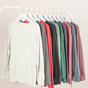 May include: A row of long-sleeved shirts in various colors, including light gray, dark gray, red, black, green, pink, blue, and mauve, hanging on white hangers. The shirts are displayed on a white clothing rack against a light pink background.