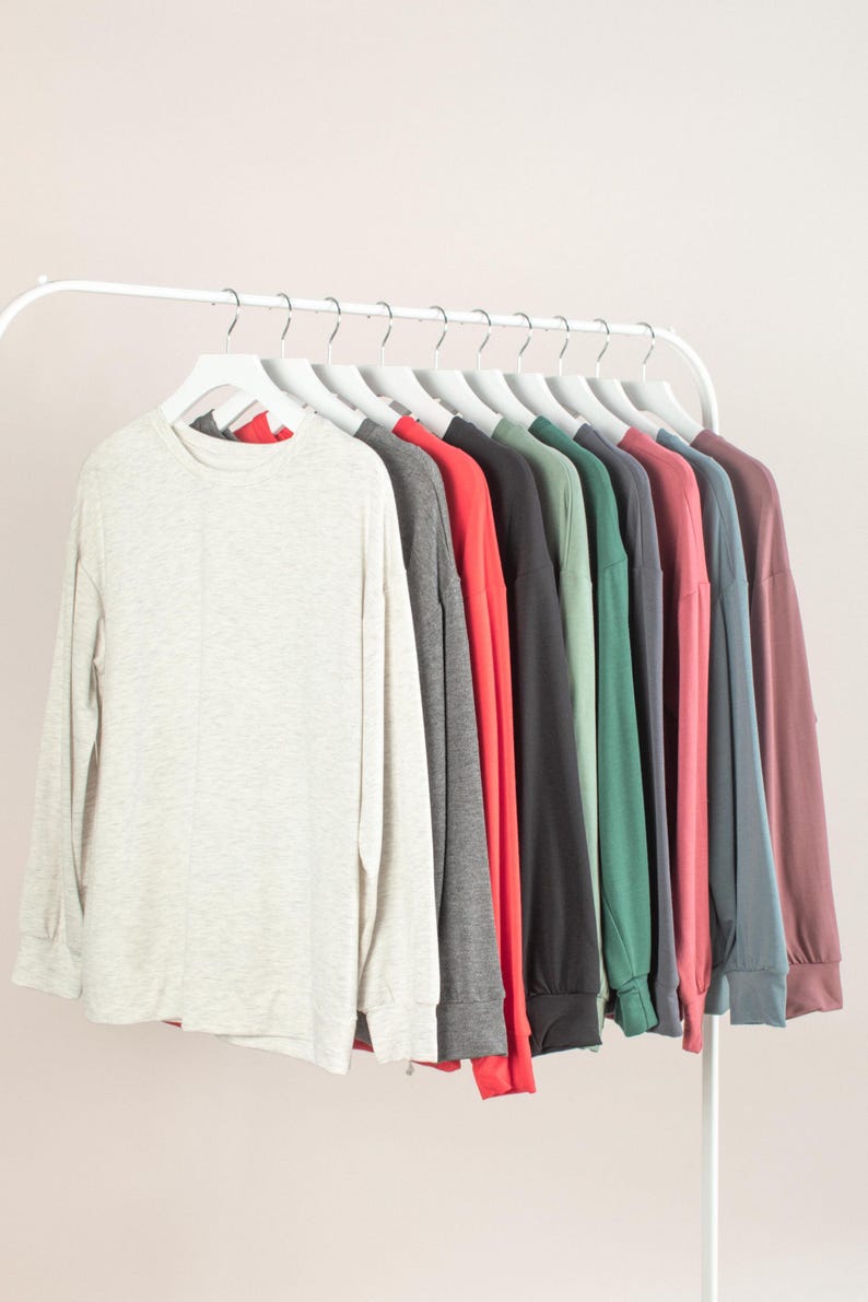 May include: A row of long-sleeved shirts in various colors, including light gray, dark gray, red, black, green, pink, blue, and mauve, hanging on white hangers. The shirts are displayed on a white clothing rack against a light pink background.