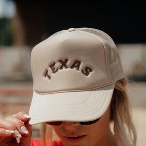 May include: A beige trucker hat with a brown embroidered "TEXAS" logo. The hat has a mesh back and a curved brim.